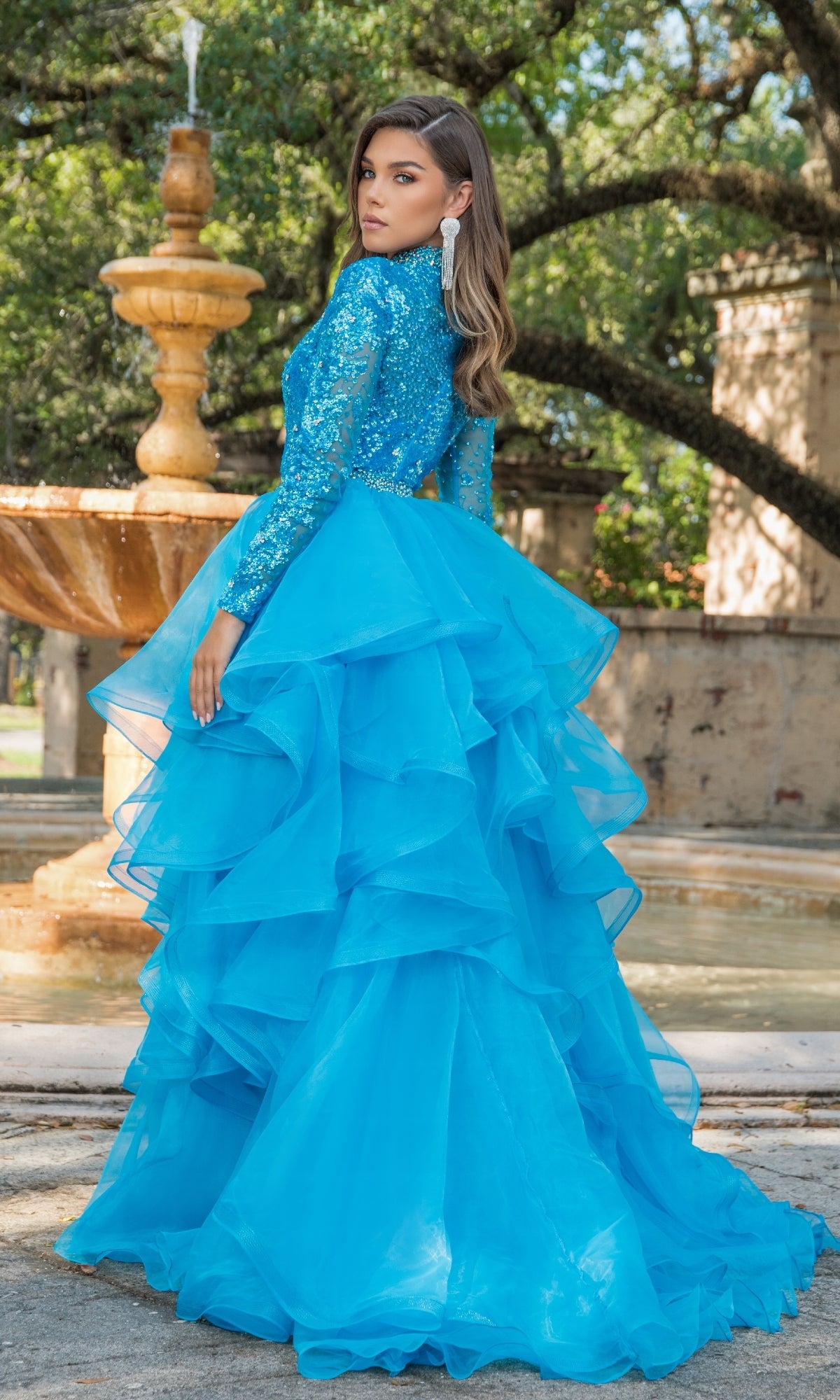 A woman stands outdoors by a stone fountain and lush greenery, wearing the Ava Presley 28595 Ruffle Short Sequin Romper in bright blue with long sleeves and a dramatic ruffled overskirt.